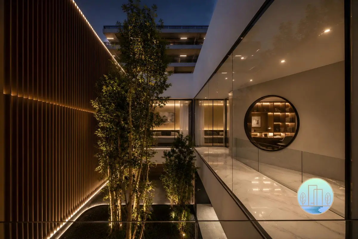 Modern luxury interior courtyard atrium at night featuring vertical wooden slats, illuminated trees, glass railings, and a circular window revealing an illuminated bookshelf.