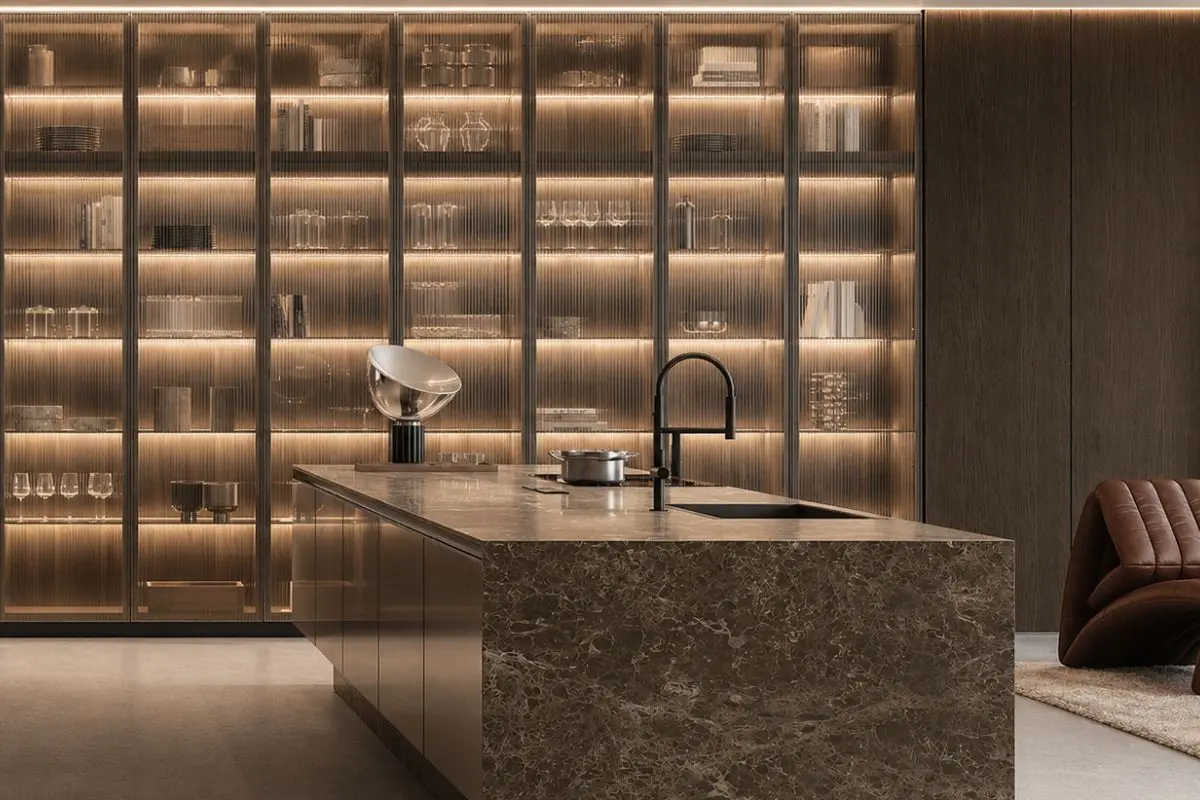 Modern kitchen featuring a large dark brown marbled island with an integrated sink and a wall of illuminated, fluted glass display cabinets filled with glassware and books.